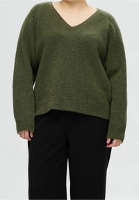 Maglione verde a scollo a V realizzato in materiale morbido e texture, con spalle scese, vestibilità ampia e polsini a coste, abbinato a pantaloni neri.