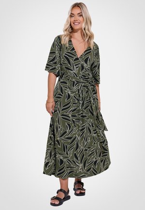 Blonde Frau trägt ein grünes Wickelkleid mit schwarzem und weißem Blattmuster, steht in schwarzen Sandalen mit Schnallen vor weißem Hintergrund.