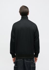 Μαύρο μπουφάν με φερμουάρ και ψηλό γιακά, κατασκευασμένο από υφή υφάσματος, με ελαστικά μανσέτες και τελείωμα. Εμφανίζεται η πίσω όψη, χωρίς σχέδια ή διακοσμητικά στοιχεία.