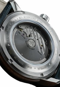 Stainless steel wristwatch back with visible movement and engravings. Features a glass case back, metallic texture, and gold accents.