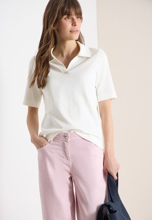 Frau trägt ein weißes Hemd mit kurzen Ärmeln und Kragen sowie hellrosa Hosen, hält eine dunkelblaue Tasche und hat eine Hand in der Hosentasche.