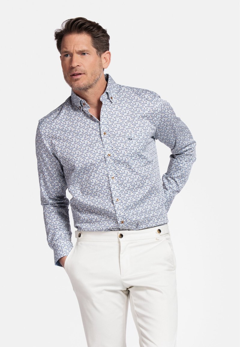 Chemise à boutons en bleu clair avec un motif géométrique, manches longues et boutons dorés contrastants. Portée avec un pantalon blanc.