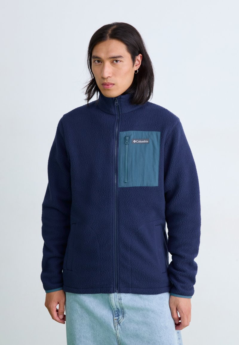 Men's navy fleece jacket with a teal zippered chest pocket, high collar, front pockets, and textured surface.