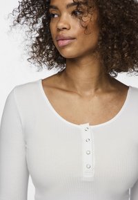 Bílý žebrovaný top s dlouhým rukávem, který má kulatý výstřih a pět stříbrných patentek vpředu, s měkkou a přiléhavou texturou.