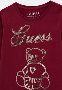Camicia bordeaux Guess con testo "Guess" in paillettes argentate e un orsetto che indossa una gonna con il logo sul davanti.
