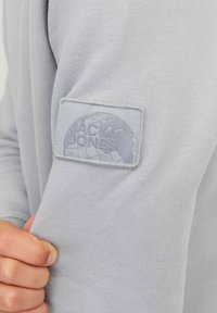 Svjetlosiva dukserica mekane teksture, sivi gumeni logo "JACK & JONES" na lijevom rukavu.