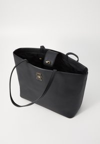 Black leather tote bag with smooth texture, featuring a gold logo accent and a top closure. Slim handles for carrying. Interior visible.