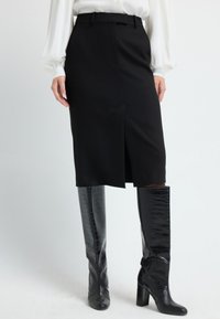 Jupe crayon noire au design ajusté, avec une fente centrale et des passants de ceinture, associée à des bottes noires en cuir croco brillantes et hautes.