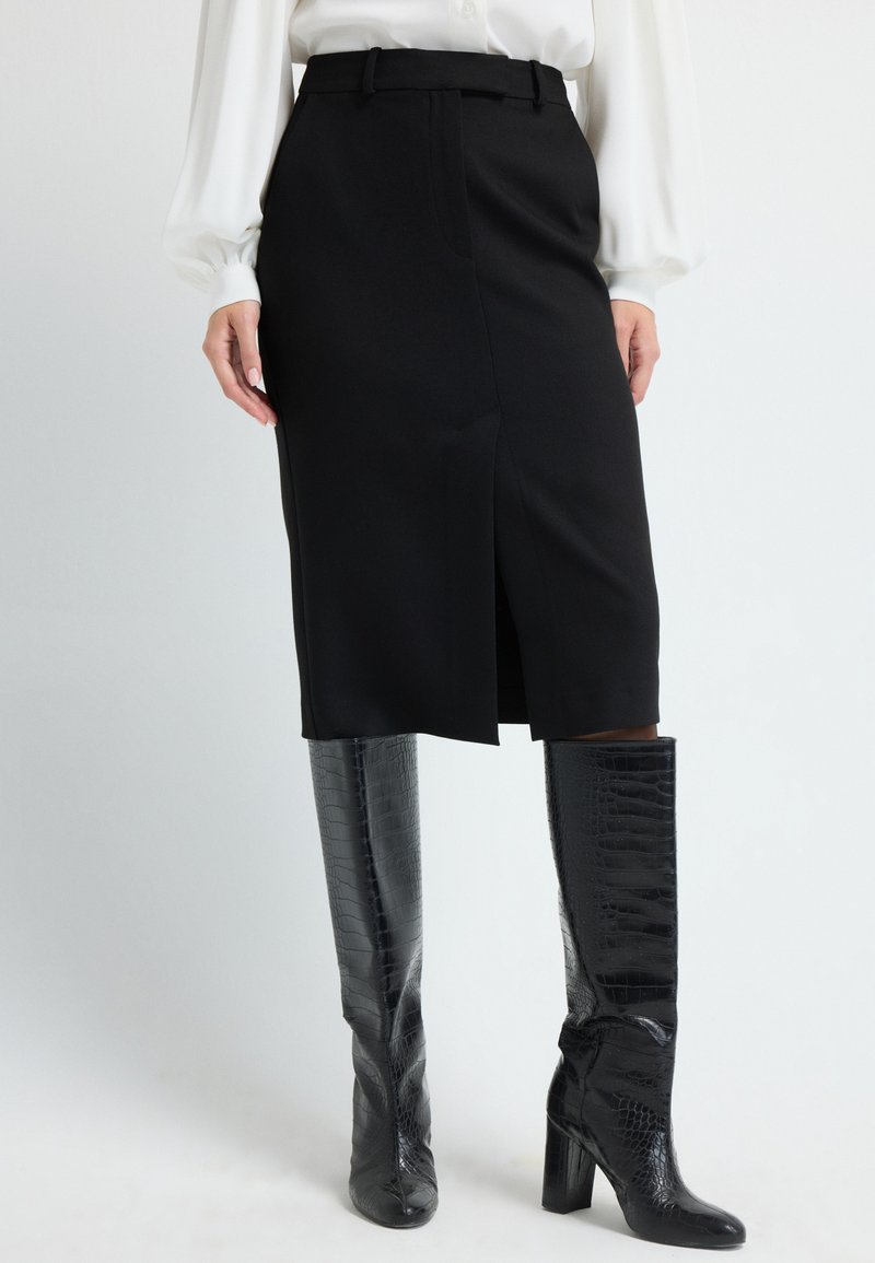 Jupe crayon noire au design ajusté, avec une fente centrale et des passants de ceinture, associée à des bottes noires en cuir croco brillantes et hautes.