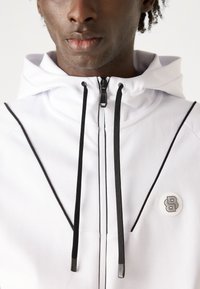 Haut de sweat à capuche zippé blanc avec des accents noirs, présentant un col haut, des coutures contrastées et un logo de marque circulaire sur la poitrine.