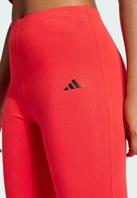 Röda träningsleggings i elastisk bomull, med en svart Adidas-logotyp nära midjan och en slät, åtsittande design.