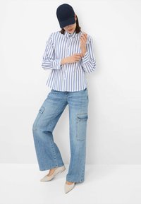 Overhemd met blauw-witte verticale strepen, gecombineerd met lichtblauwe wijde cargobroek. Beige puntige flats en een marineblauwe pet.