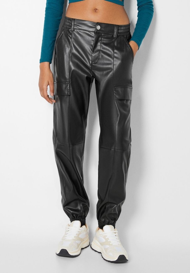 Pantalons cargo en simili cuir noir avec des poignets élastiques, deux poches avant et une fermeture à bouton, associés à des baskets blanches.