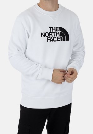 Mann trägt weißen Pullover mit schwarzem "THE NORTH FACE" Logo, kombiniert mit schwarzen Cargohosen, steht vor einfachem Hintergrund.