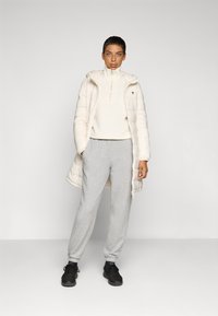 Pufferjack in licht beige, met een ritssluiting; witte hoge nektrui; grijze joggingbroek; zwarte sportschoenen; gladde texturen.
