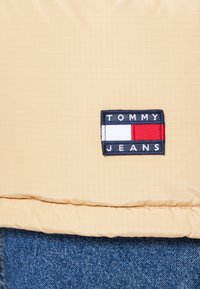 Gelbe Steppjacke mit einem Gittermuster; ausgestattet mit einem dunkelblauen rechteckigen Patch mit "TOMMY JEANS" und drei farbigen Streifen in Rot, Weiß und Blau.