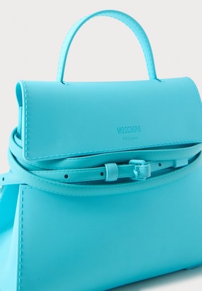 Μπλε τσάντα Moschino Milano με επάνω λαβή και λεπτή ρυθμιζόμενη ζώνη σε απλό ανοιχτό φόντο.