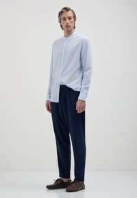 Camicia button-up azzurra chiara con colletto a band, maniche lunghe, abbinata a pantaloni elasticizzati blu scuro in vita e scarpe marroni con lacci.