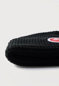 Μαύρο πλεκτό σκουφάκι με ριμπ ύφανση, διακοσμημένο με στρογγυλό κόκκινο και λευκό λογότυπο. Η διπλωμένη άκρη προσθέτει στο συνολικό του σχεδιασμό.