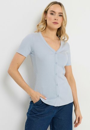 Frau mit blondem Haar, die ein hellblaues kurzärmeliges, geknöpftes Oberteil und dunkelblaue Hose trägt, die mit einer Hand in der Tasche steht.