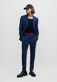 Marineblauer Anzug mit einem figurbetonten schwarzen Tanktop, rotem Markengummizug und schwarzen Sneakers. Das Model trägt eine schwarze Mütze und hat gerade geschnittene Hosen.