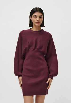 Femme aux cheveux raides et foncés vêtue d'une robe en maille côtelée bordeaux à manches longues et taille ajustée, debout devant un fond uni.