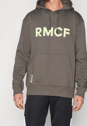 Personne portant un sweat à capuche marron avec de grandes lettres "RMCF" jaune clair à l'avant et une poche kangourou, assorti à un pantalon noir.