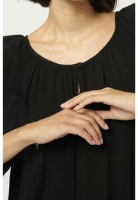 Blouse noire texturée avec un col rond et un détail de bouton à l'avant, accompagnée de mains fines portant un délicat bracelet en argent.