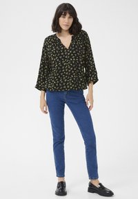 Blouse noire avec un motif floral jaune, décolleté en V, manches cloche trois-quarts. Associée à un jean bleu slim et des mocassins noirs.