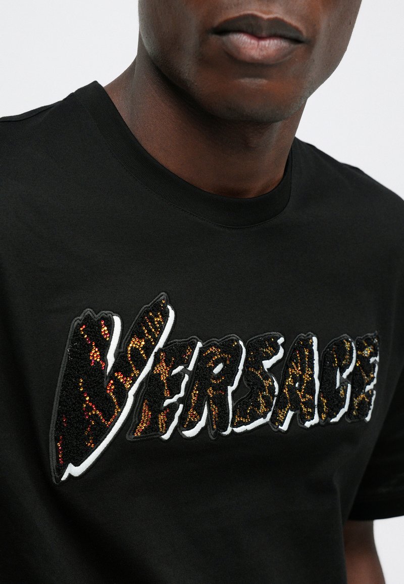 T-shirt en coton noir avec un logo "VERSACE" brodé texturé et multicolore, accentué par des perles brillantes délimitant les lettres.