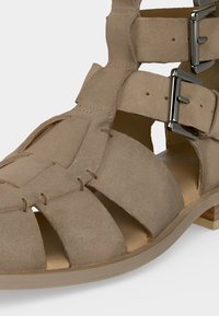 Μπεζ σουέτ σανδάλι σε στυλ μονομάχου με πολλαπλά λουράκια, δύο ασημένια αγκράφες, ανοιχτά δάχτυλα και χαμηλό ξύλινο τακούνι.