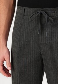 Close-up van een persoon die donkergrijze strepenbroek draagt met een zwarte trekkoord dat in een strik op de taille is geknoopt, hand rustend nabij de zijkant.