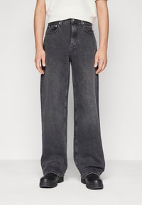 Jean en denim noir à jambes larges avec une texture délavée, taille haute, poches avant et fermeture à bouton, assorti à des chaussures noires.