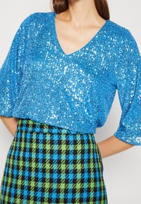 Top blu con paillettes, scollatura a V e maniche corte a sbuffo, abbinato a una gonna a quadri verde e nera. Superficie testurizzata e lucente.