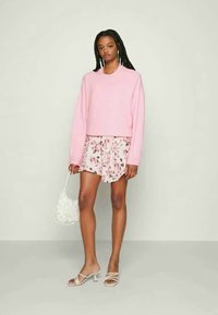 Sweater rosa, cropped, com mangas longas, combinado com uns calções com padrão floral. A modelo segura uma mala branca, texturizada, e usa sandálias de salto transparente.