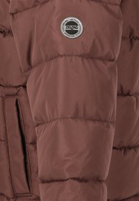 Veste matelassée marron avec un design cousu, dotée d'un patch logo métallique rond et d'une poche latérale, fabriquée en matériau lisse et glossy.