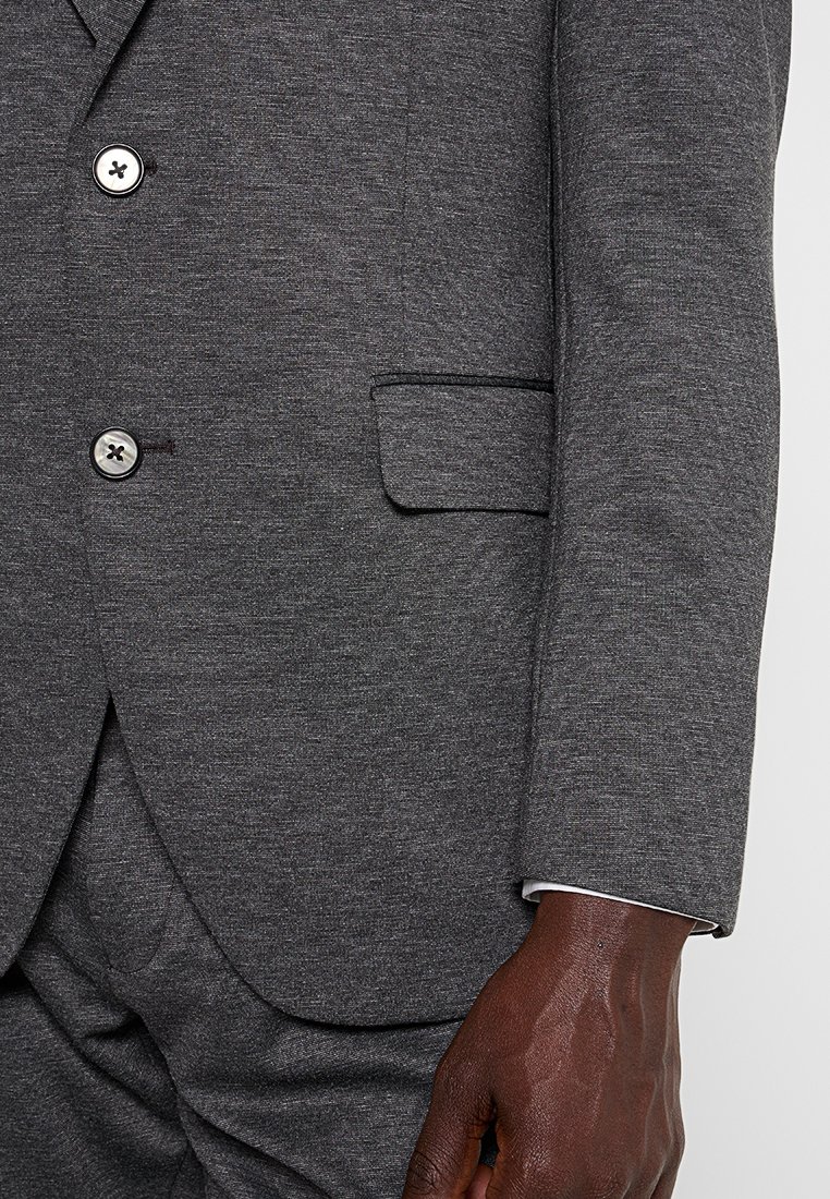 Chaqueta de traje gris oscuro con dos botones y un bolsillo con solapa, llevada sobre pantalones a juego, mostrando una mano de piel oscura descansando a un lado.