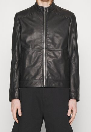 Person iført en tætsiddende sort læderjakke med lynlås foran, stående med armene afslappet ned langs siderne mod en ensfarvet lys baggrund.