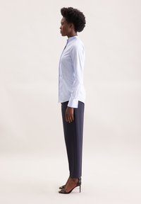 Camicia lunga a maniche con colletto rialzato, a righe blu chiaro, indossata con pantaloni scuri e tacchi neri. Tessuto liscio, vestibilità su misura. Vista laterale.