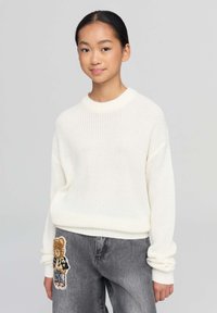 Biały sweter z dzianiny o okrągłym dekolcie, w zestawieniu z szarymi dżinsami z łatą w kształcie misia, prezentujący swobodny, warstwowy wygląd.