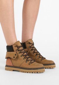 Kožené turistické boty v opálené barvě s černou kotníkovou podporou, rezavými akcenty, kovovými očky a robustní, strukturovanou podrážkou. Design na šněrování.