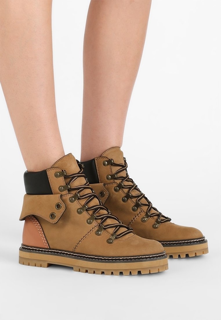 Kožené turistické boty v opálené barvě s černou kotníkovou podporou, rezavými akcenty, kovovými očky a robustní, strukturovanou podrážkou. Design na šněrování.