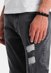 Mano de hombre con pulsera de cuentas negras al lado de jeans oscuros rasgados y camisa blanca contra un fondo liso.