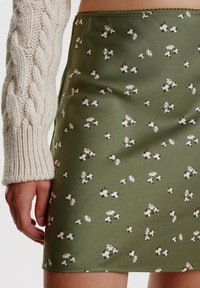 Vihreä kukallinen mini-hame, jossa on valkoisia ja keltaisia kukkakuoseja. Silkkinen kangas, jossa on istuva muotoilu ja reunassa aallotettu helma.