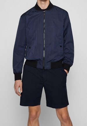 Mand iført en mørkeblå bomberjakke med sorte ribkanter ved ærmer og kant, kombineret med matchende mørkeblå shorts, højre hånd i lommen.