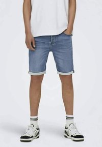 Gaiši zilie džinsu šorti ar sarullētām malām, piedāvājot klasisko piecu kabatu dizainu. Savienoti ar baltām kedām un svītrainām zeķēm.