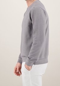 Felpa grigia con maniche lunghe, scollatura rotonda e texture morbida. Presenta una vestibilità comoda e polsini a coste, indossata con pantaloni bianchi.