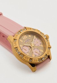 Ručno sat s ružičastim silikonskim remenom, kućištem zlatne boje, sjajnom lunetom i teksturiranim zlatnim brojčanikom s ružičastim podbrojčanicima i izraženim oznakama sati.