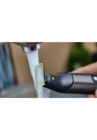 Elektrische scheermes met een zwarte behuizing en gele accenten. Close-up van het mes onder stromend water, waarbij de gestructureerde snijkant wordt getoond.