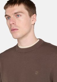 Brun tröja med rund halsringning och ribbade ärmmuddar. Har en liten, broderad logotyp i en ljusare nyans. Mjuk, texturerad stickad material.